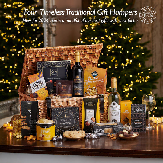 the blog cover to the latest blog from Spicers of Hythe on luxury traditional gift hampers. The image features an open hamper basket bursting with prosecco, wine, crisps & other luxury artisan goods. It is sat on a white and silver table top.